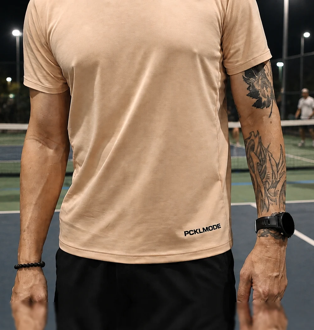PCKLMODE performance tee in sand on a lit court at night with a rally behind, the mark embroidered at the right hem